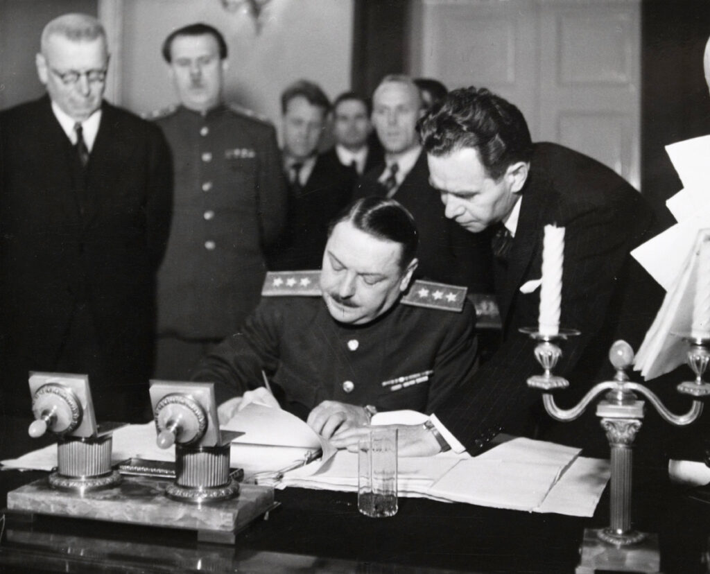Mustavalkoinen valokuva Valtioneuvoston juhlahuoneistosta. Kuvan keskellä, kirjoituspöydän ääressä, istuu kenraali Andrei Ždanov allekirjoittamassa sopimusta. Hänen vierellään on mies, joka on kumartunut pitelemään sopimuksen käännettyä lehteä sileänä. Taustalla vasemmalla, hieman kauempana, seisoo Suomen silloinen pääministeri J.K. Paasikivi. Kuvan etualalla on työpöydällä olevia tavaroita: mustepullot, juomalasi ja koristeellinen kynttilänjalka.