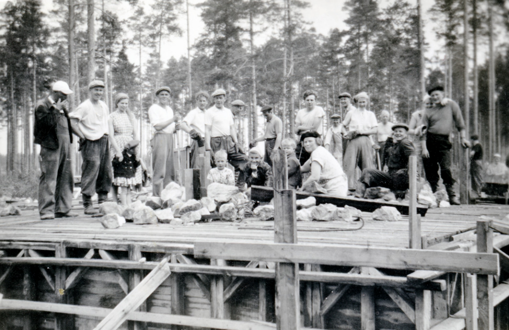 Ryhmäkuva rintamamiestalon rakennustalkoista. Kellari on saatu valettua ja talkooporukka poseeraa kameralle vielä seinättömän ensimmäisen kerroksen laudoituksilla.
