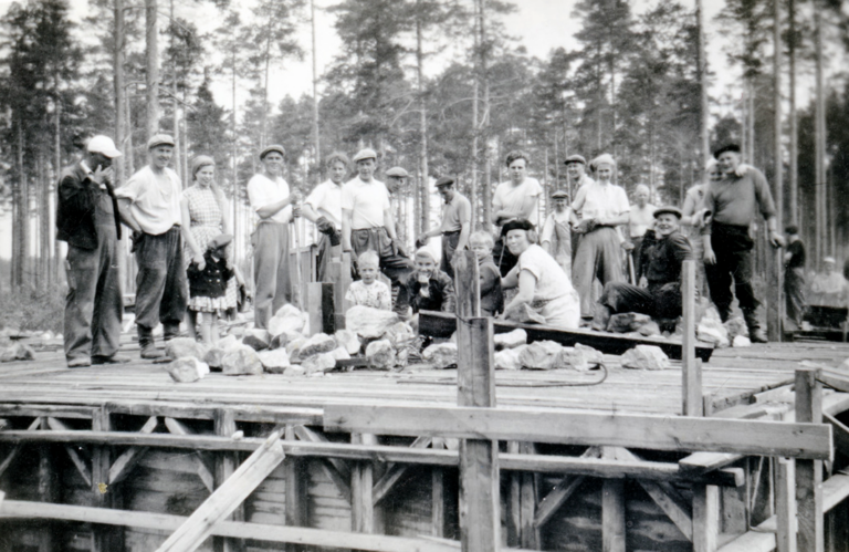 Ryhmäkuva rintamamiestalon rakennustalkoista. Kellari on saatu valettua ja talkooporukka poseeraa kameralle vielä seinättömän ensimmäisen kerroksen laudoituksilla.