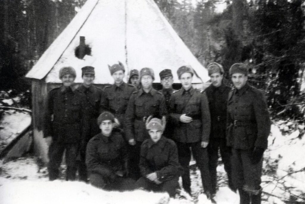 Mustavalkoinen valokuva. Joukko suomenjuutalaisia sotilaita poseeraa yhteiskuvassa pienen kenttäsynagogan edessä. Sotilaat valmistivat synagogan pahvista ja vanerista. Sisällä oli kamiina, jonka savupiippu törröttää kuvassa ulos katossa olevasta reiästä.