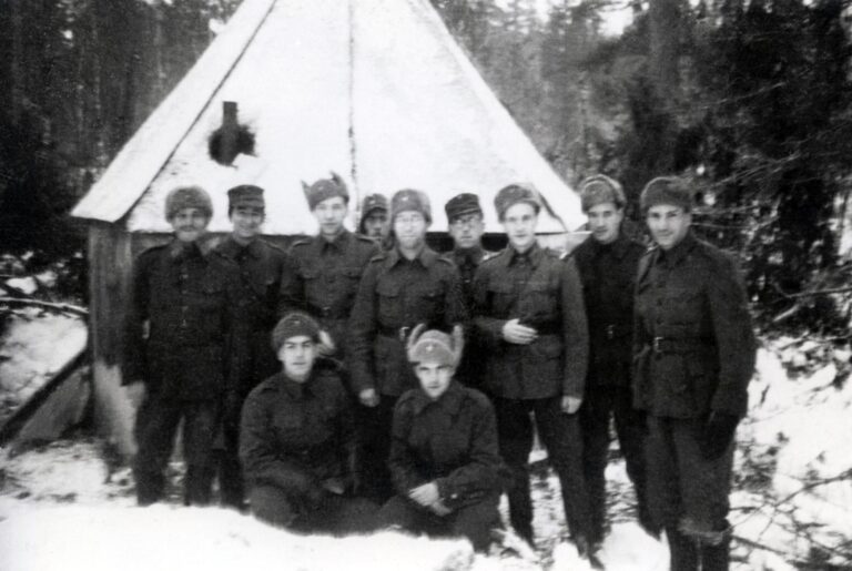 Mustavalkoinen valokuva. Joukko suomenjuutalaisia sotilaita poseeraa yhteiskuvassa pienen kenttäsynagogan edessä. Sotilaat valmistivat synagogan pahvista ja vanerista. Sisällä oli kamiina, jonka savupiippu törröttää kuvassa ulos katossa olevasta reiästä.