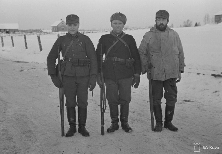 Mustavalkoinen valokuva. Kolme suomalaista sotilasta seisoo rinnatusten kohti kameraa. Kullakin on hieman erilainen univormu. Ensimmäisellä miehellä on sarkaunivormu ja kenttälakki. Toisella on yllään takki tummempaa kangasta. Kolmannella taas on vaaliea hupullinen takki. Taustalla on talvista maalaismaisemaa.