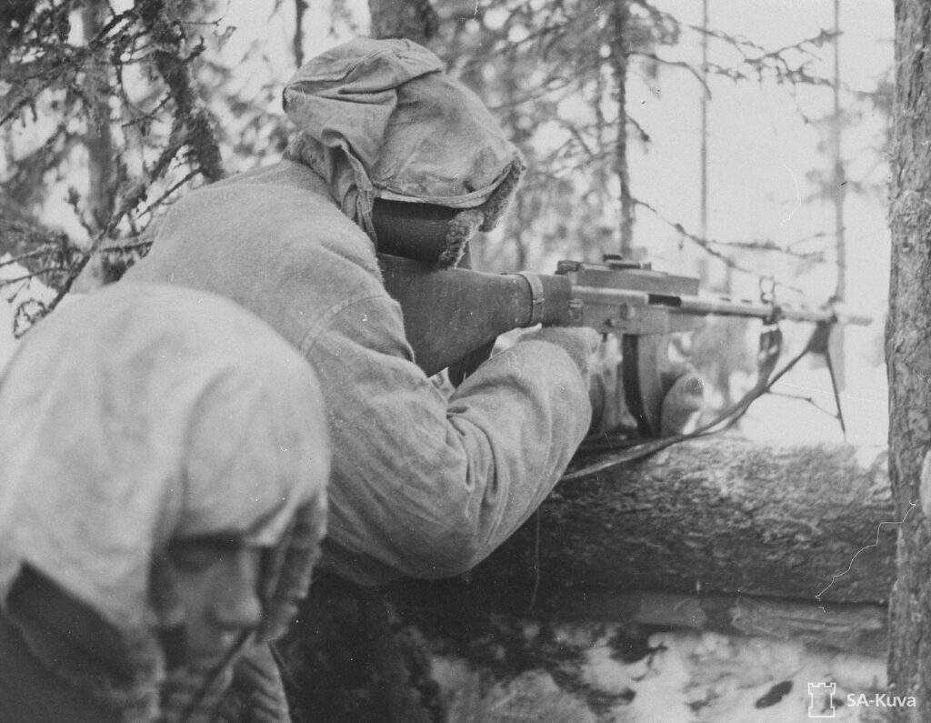Mustavalkoinen valokuva. Kuvassa on kameraa kohti selin sotilas, joka tähtää pikakiväärillä juoksuhaudan reunan yli. Kuvan alareunassa vasemmalla näkyy toisen sotilaan kasvot. Molemmilla on yllään vaaleat lumipuvut, huppu päähän nostettuna.