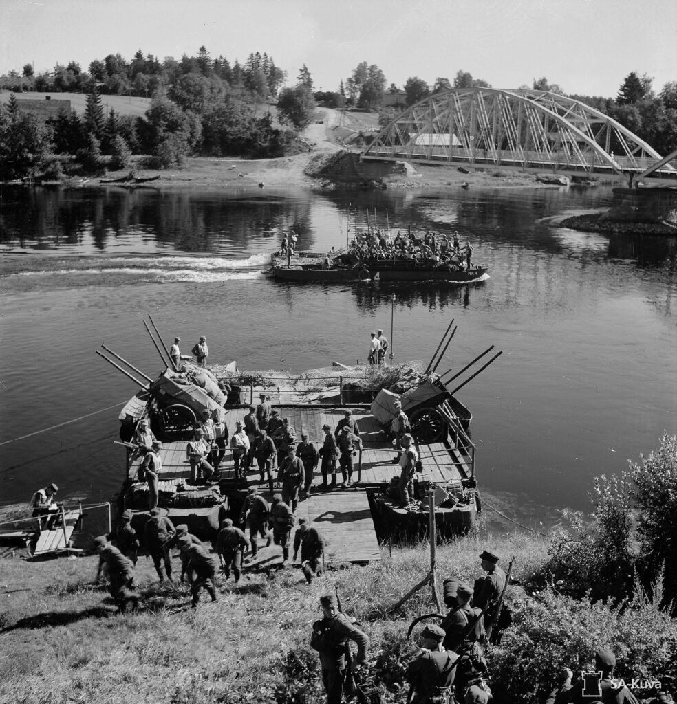 Mustavalkoinen valokuva. Joukko sotilaita on nousemassa maihin neliön muotoiselta lautalta. Lautta on kuljettanut joen yli myös kärryjä, jotka on lastattu tavaralla ja peitetty kankailla. Taustalla näkyy toinen vedessä liikkuva lautta sekä joen yli kulkeva silta.