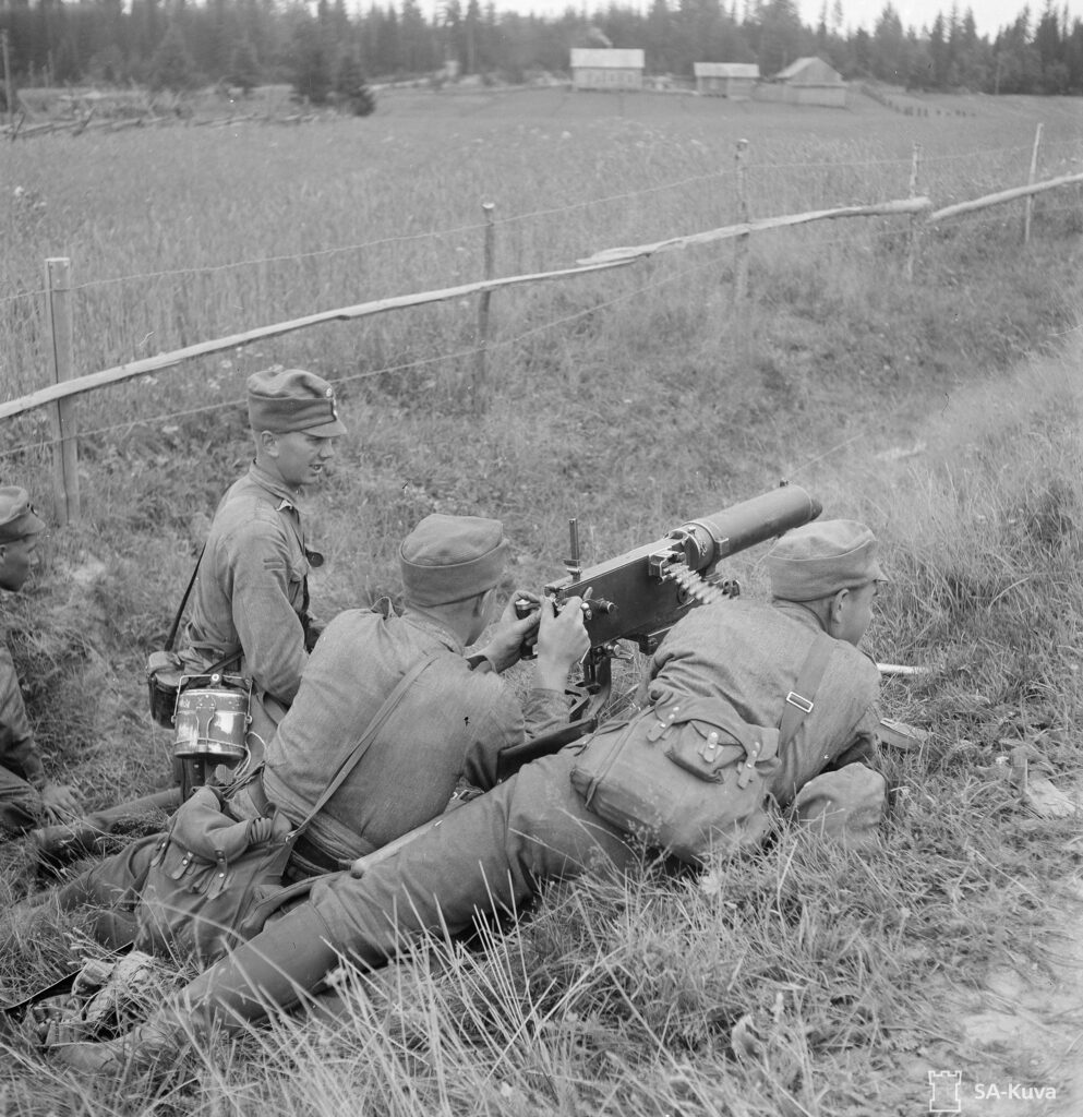 Mustavalkoinen valokuva. Kolme suomalaista sotilasta harjoittelee konekiväärillä ampumista pellon reunalla. Yksi sotilaista istuu kyykyssä ja tarkkailee kahden maassa makaavan toverinsa toimintaa.