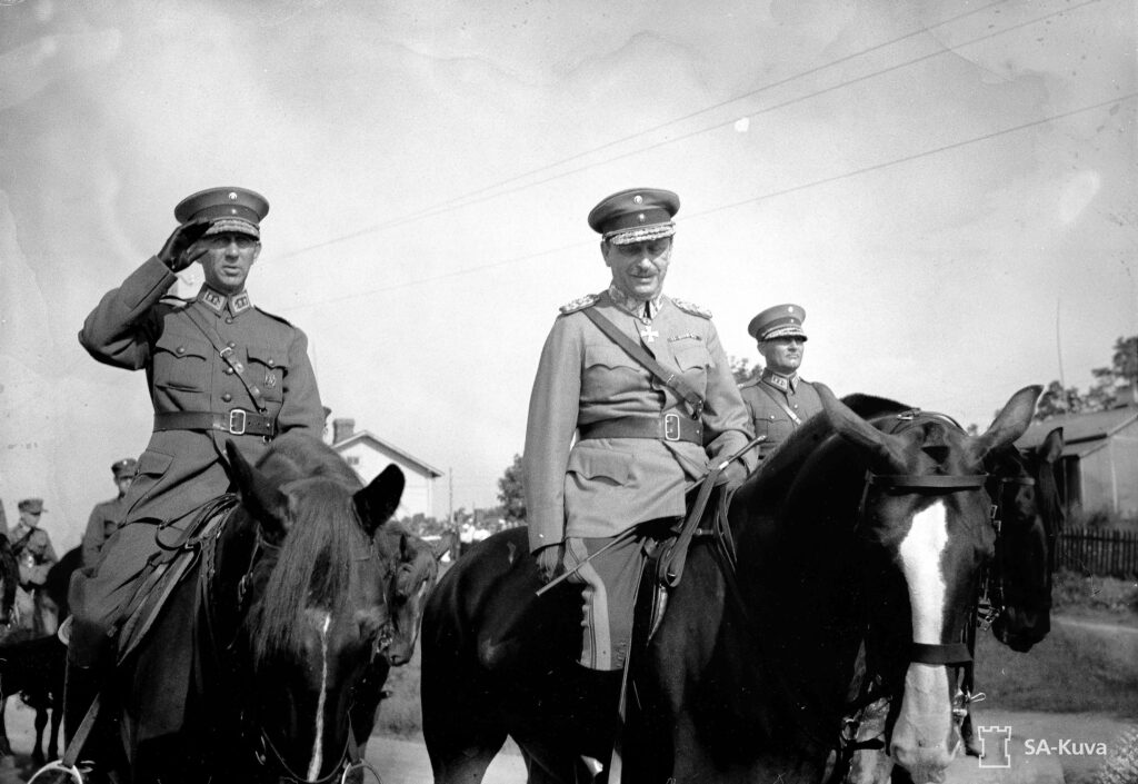 Mustavalkoinen valokuva. Mannerheim on kuvattu univormussaan hevosensa selässä. Hänen rinnallaan on ratsain kaksi upseeria, joista toinen on kohottanut kätensä tervehdystä tai kunnian tekemistä varten.